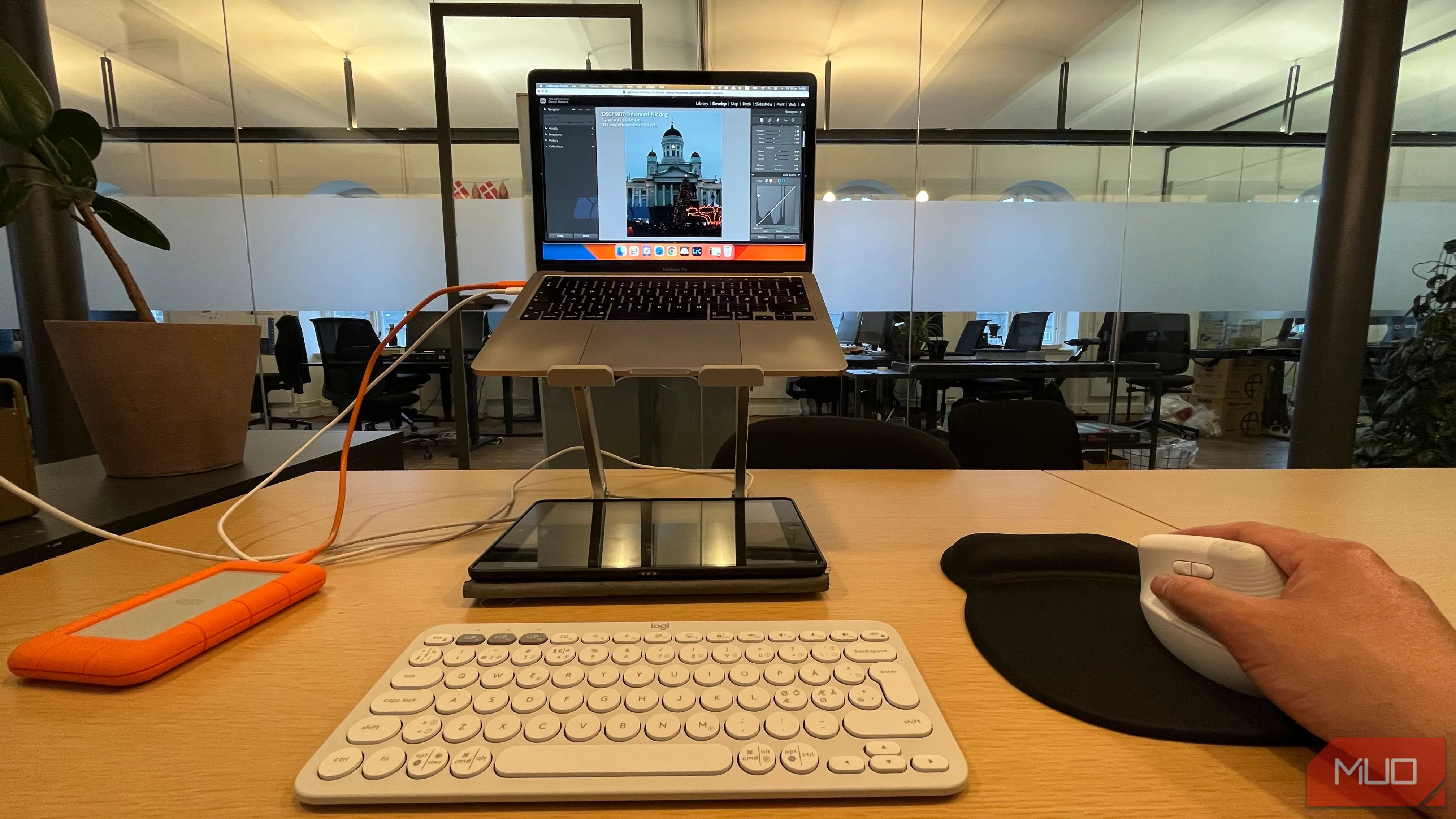 Người dùng đang chỉnh sửa ảnh trên laptop đặt trên giá đỡ, sử dụng chuột không dây và bàn phím ngoài